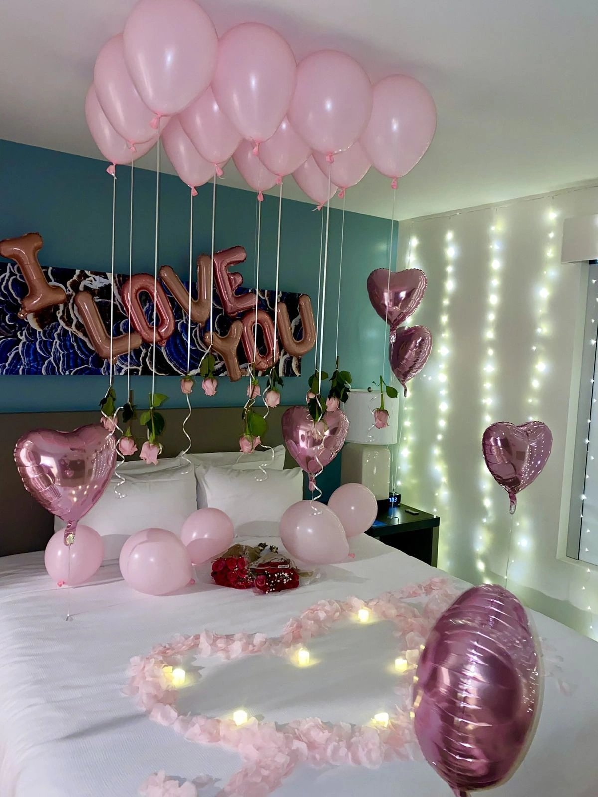 Romantic anniversary bedroom decoration with pink balloons, heart foils, LOVE YOU letters, fairy lights and a rose petal heart with candles