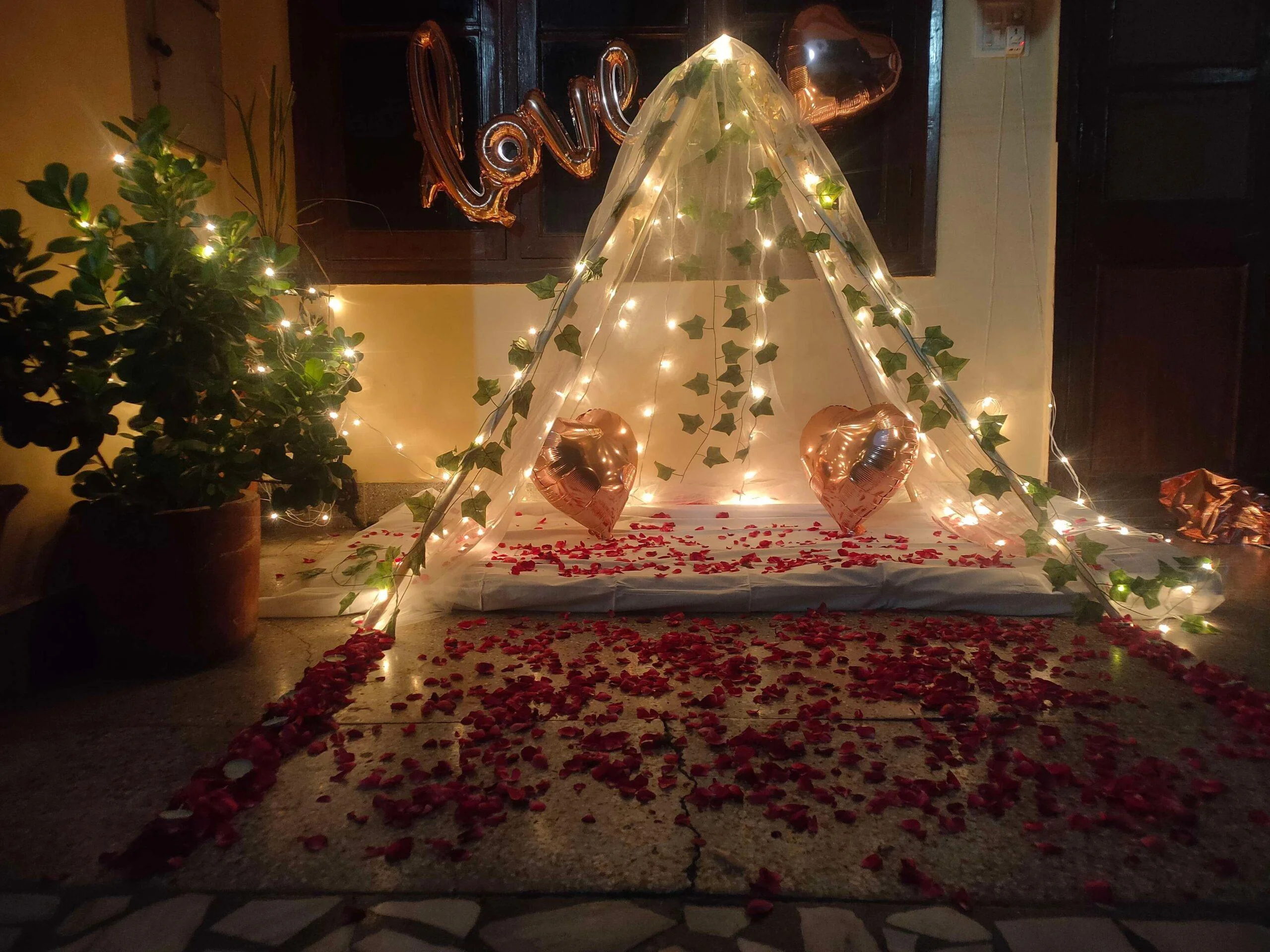 Romantic anniversary canopy decoration with fairy lights, rose petals, heart balloons and LOVE foil letters