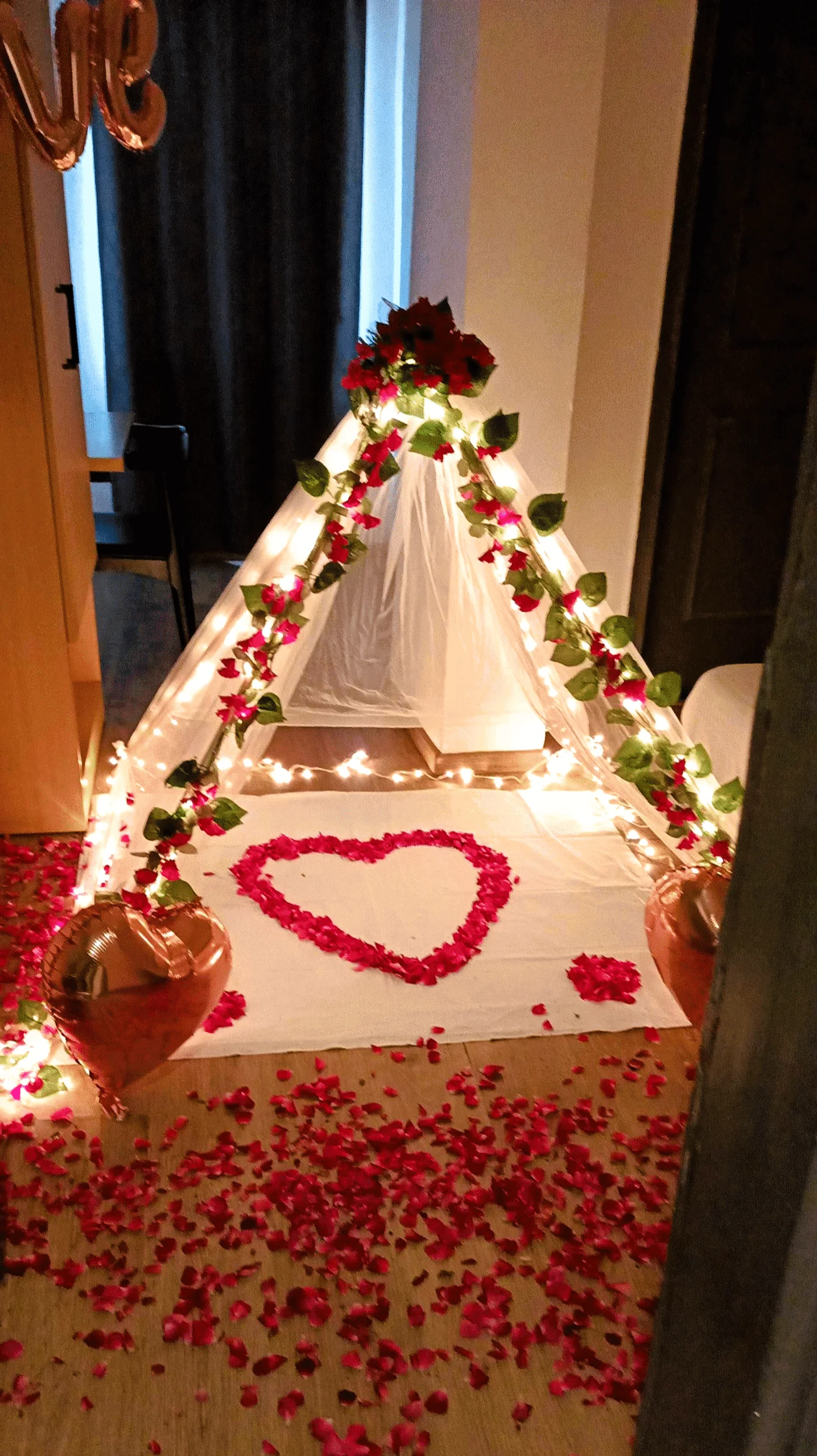 Romantic anniversary canopy decoration with fairy lights, red flowers, greenery, rose petal heart and scattered rose petals