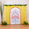 Printed floral puja backdrop framed by yellow marigold strings with two floor bouquets and an additional tall golden pot with grass