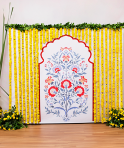 Printed floral puja backdrop framed by yellow marigold strings with two floor bouquets and an additional tall golden pot with grass