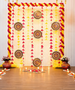 Pink and yellow Diwali backdrop with triangular tassels, patterned rangoli hangings, decorated pots and additional floor rangoli with LED candles