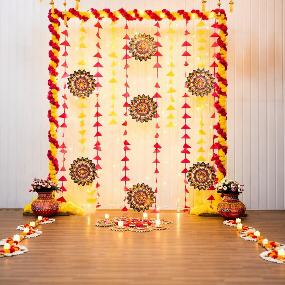 Pink and yellow Diwali backdrop with triangular tassels, patterned rangoli hangings, decorated pots and additional floor rangoli with LED candles