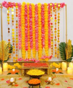 Alternative pink and yellow garland puja backdrop with gold circular hangings, palm leaves, a chowki and LED candles