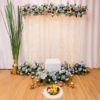 Pink, white and blue floral pooja backdrop with hanging flower strings and additional chowki, brass accessories, fruits and candle stands