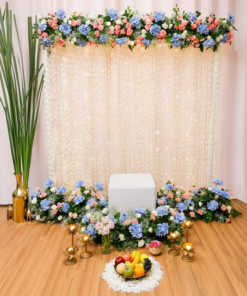 Pink, white and blue floral pooja backdrop with hanging flower strings and additional chowki, brass accessories, fruits and candle stands